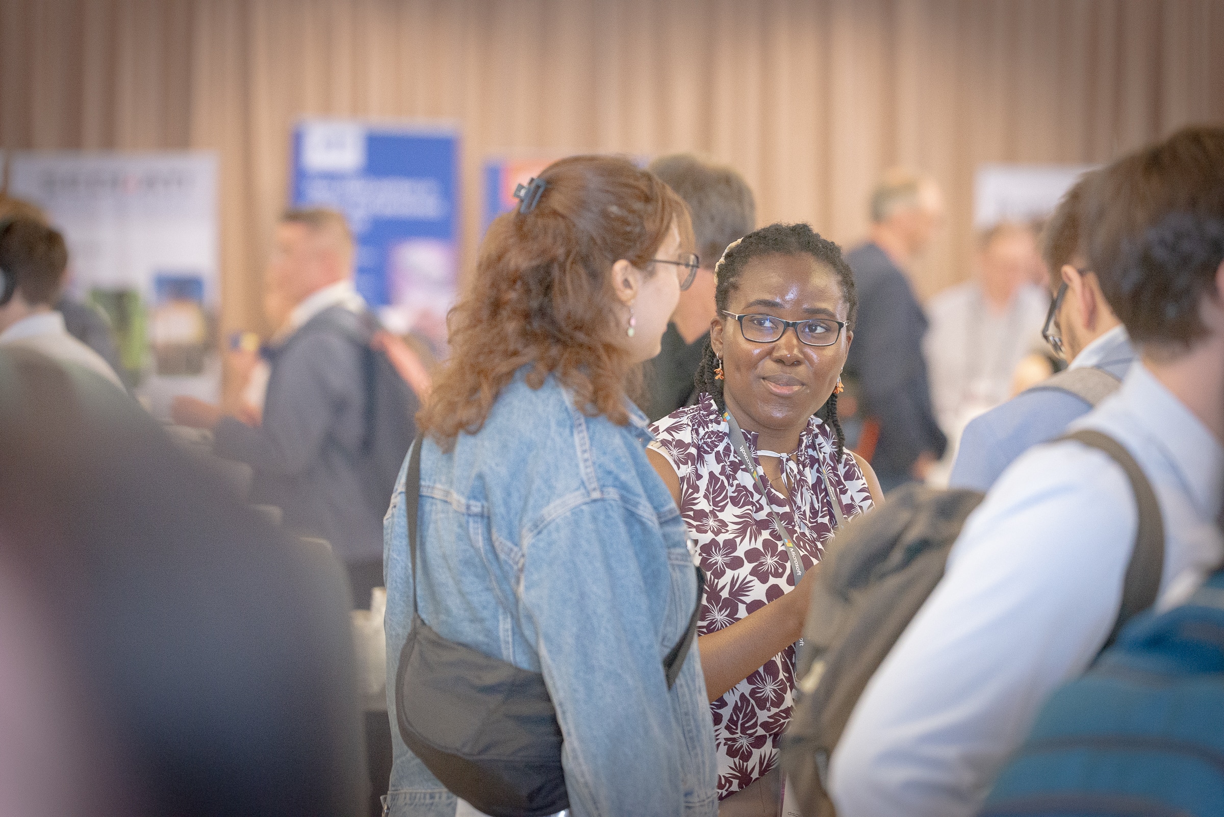 A networking event at Industry Connect: Matchmaking in Photonics, Microelectronics, and Quantum, showcasing attendees engaged in conversations. The focus is on two participants in the foreground, one wearing a floral blouse and glasses, and the other in a denim jacket, discussing or exchanging ideas. The background features event booths and blurred participants, highlighting an active and collaborative environment.