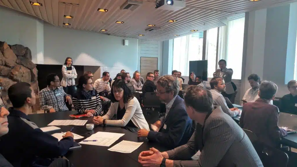 Participants engaging in discussions during the PREIN Workshop on On-Chip Photonics for Sensing held on 18 November 2024. The image captures a collaborative atmosphere, with attendees seated around tables in a well-lit conference room featuring large windows and a natural stone wall. Papers, laptops, and coffee cups are visible, indicating an interactive and productive session focused on photonics and sensing technologies.