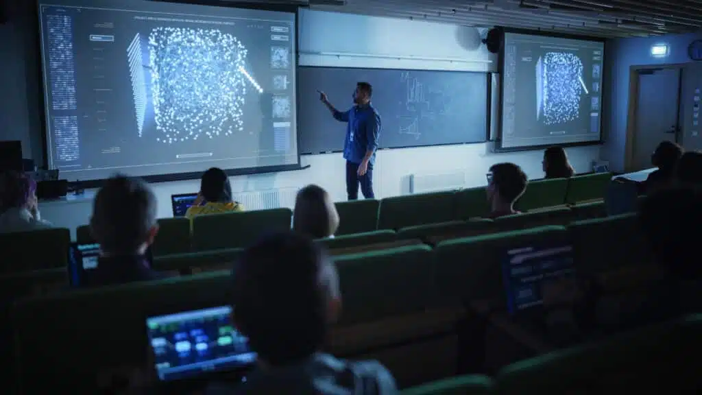 A classroom scene during the PREIN Workshop on Quantum Research & Technologies, held from 12-14 November 2024. The presenter stands at the front, pointing to a projected screen showcasing advanced scientific visualizations, likely related to quantum research or technologies. Participants sit in rows of green seats, some working on laptops displaying similar content to the presentation. The chalkboard in the background includes mathematical and technical diagrams, complementing the quantum-focused discussion.